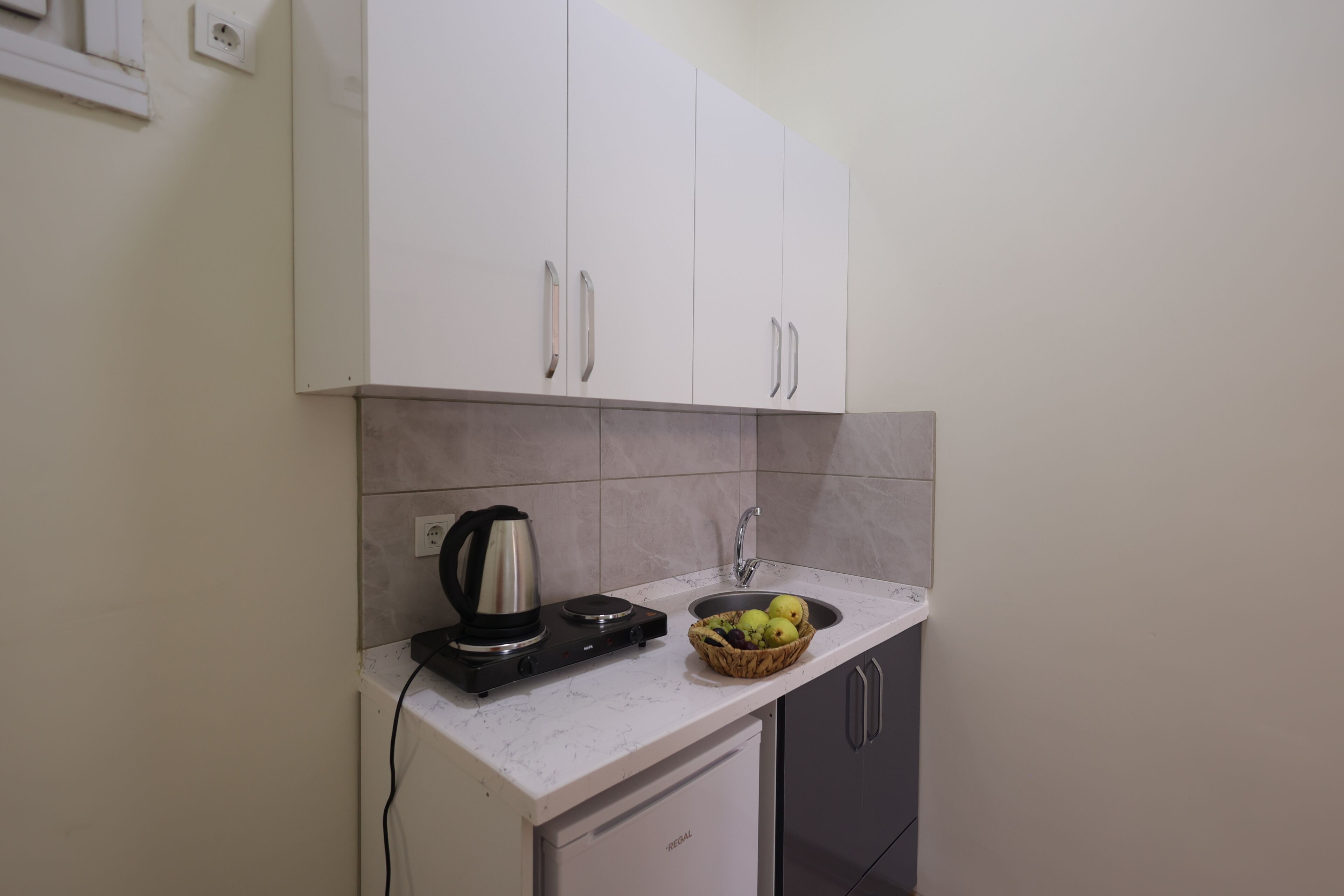 Una cocina compacta con armarios blancos, un lavamanos integrado y una encimera con hervidor y frutero.