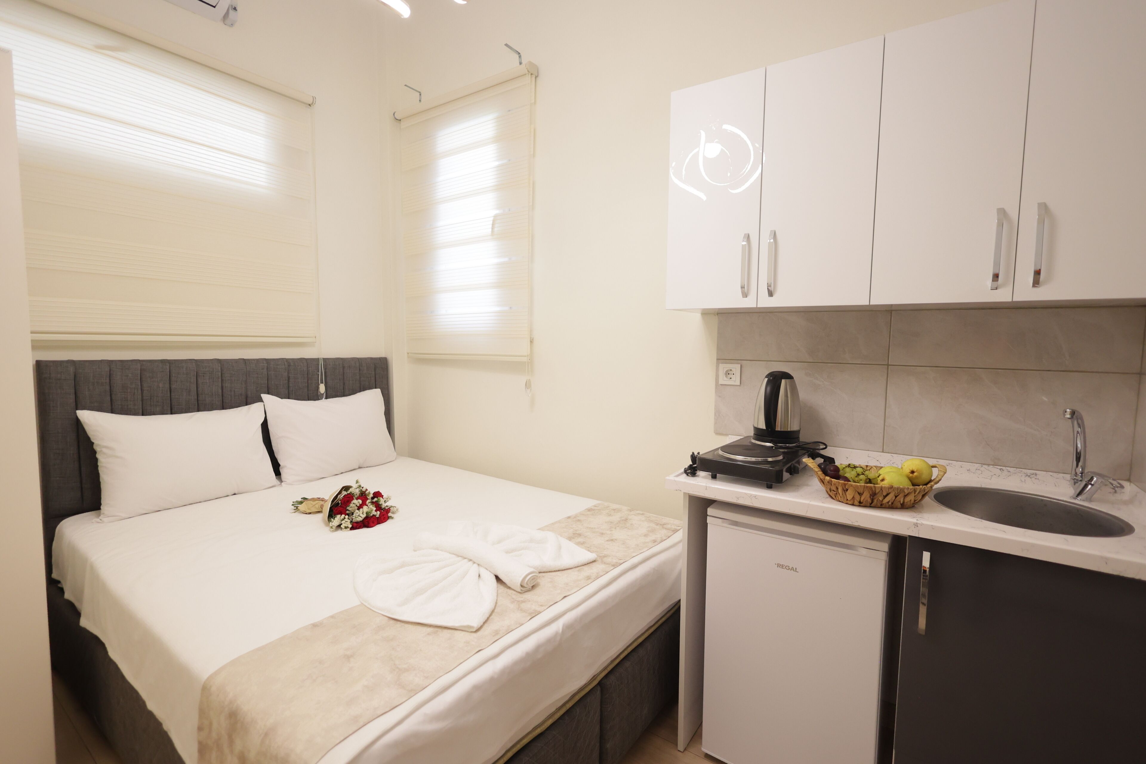 Habitació d'hotel compacta amb un llit, una cuineta petita i una finestra amb persianes.