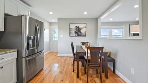 Una cocina moderna con electrodomésticos de acero inoxidable, una zona de comedor con sillas de madera y una pared gris con un cuadro enmarcado.