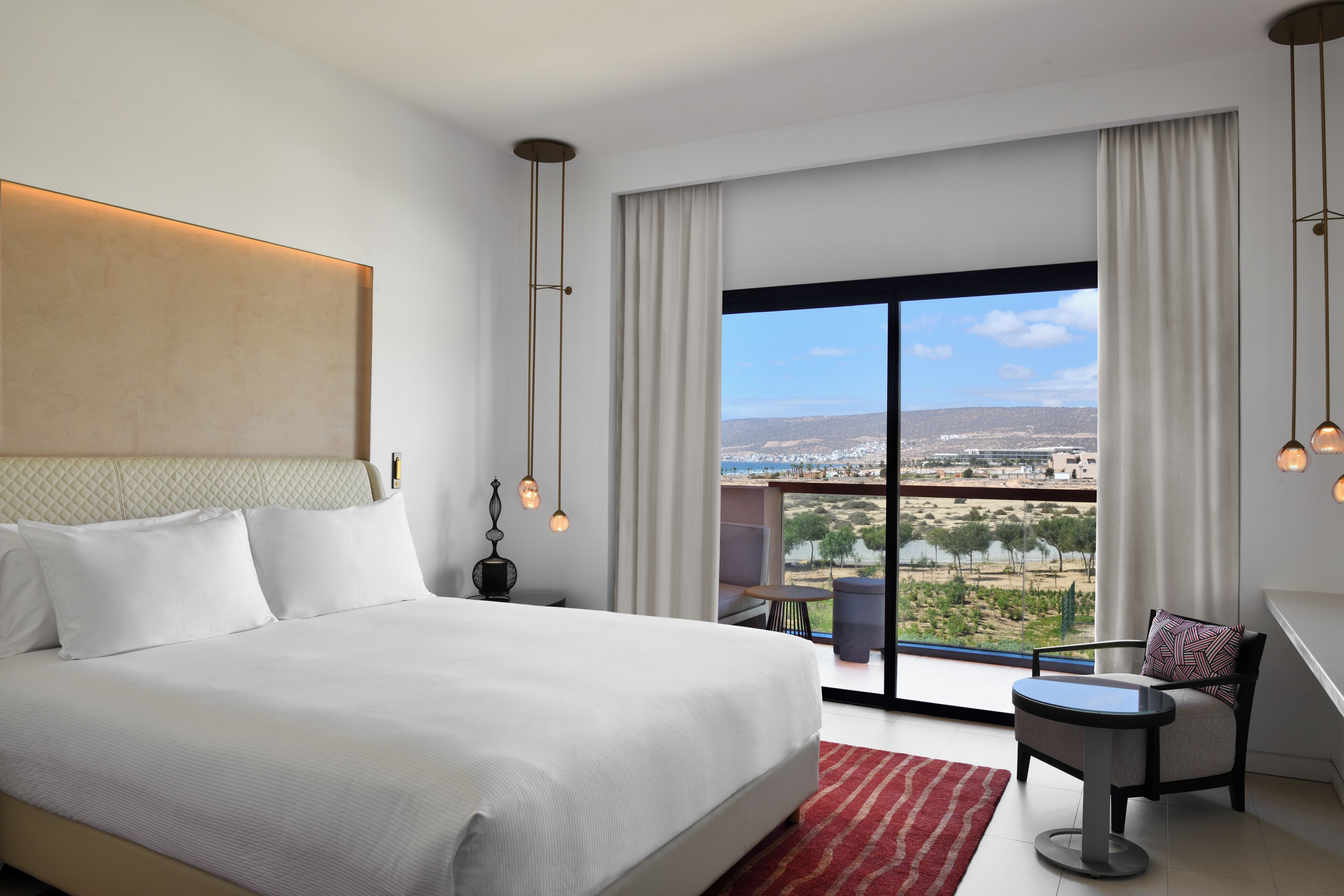 Une chambre d’hôtel moderne avec un grand lit, une petite table, une chaise et un balcon offrant une vue sur la ville.