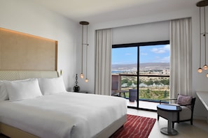 A modern hotel room with a large bed, a small table, a chair, and a balcony with a city view. - Hilton Taghazout Bay Beach Resort & Spa (Taghazout)