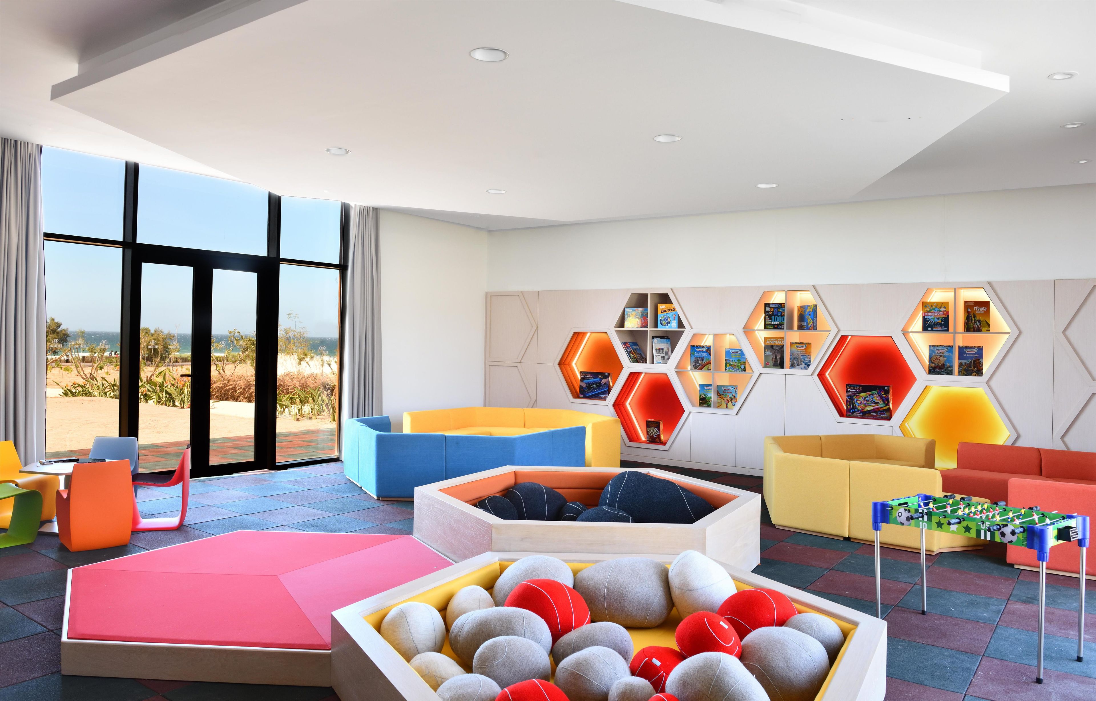 Un interior moderno con asientos coloridos, un ventanal grande y una pared con estantes hexagonales que exhiben libros.