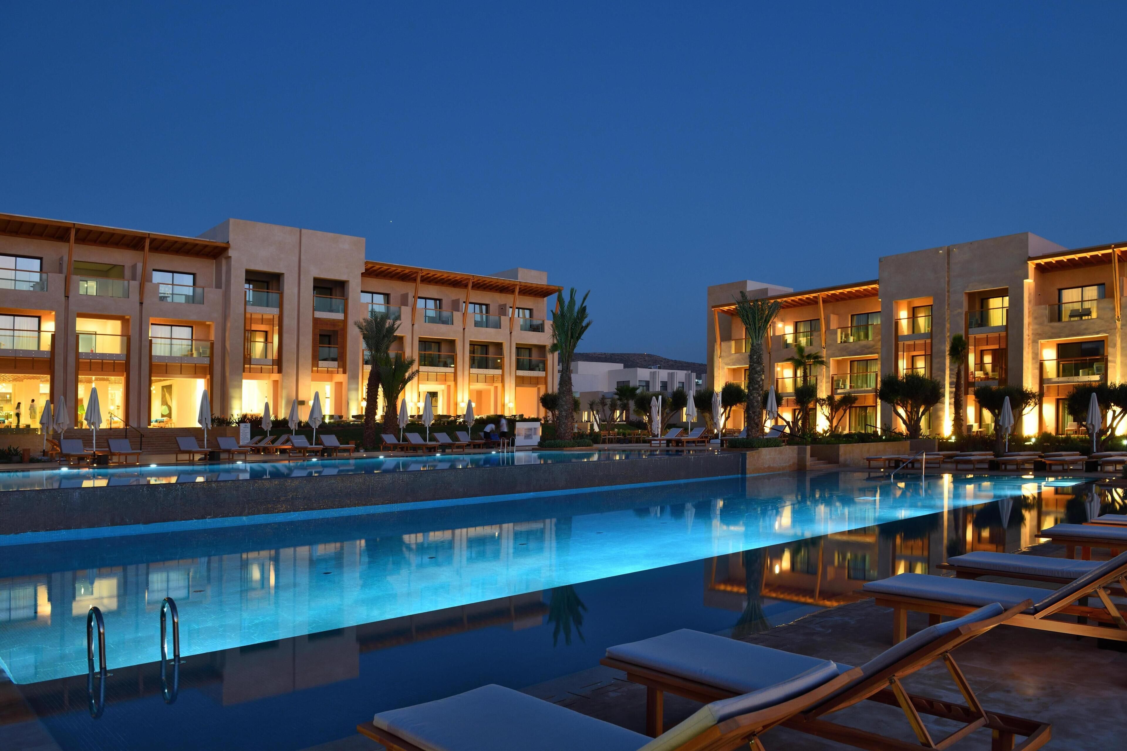 Un hôtel doté d’une piscine, de chaises longues et d’une façade bien éclairée.