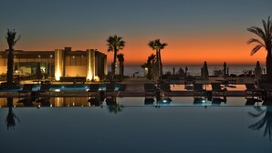 Zona de piscina de un hotel con sillones reclinables, palmeras y vistas al océano al atardecer.