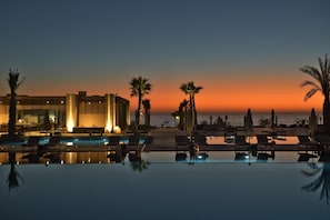 Un espace piscine d’hôtel avec des chaises longues, des palmiers et une vue sur l’océan au coucher du soleil.
