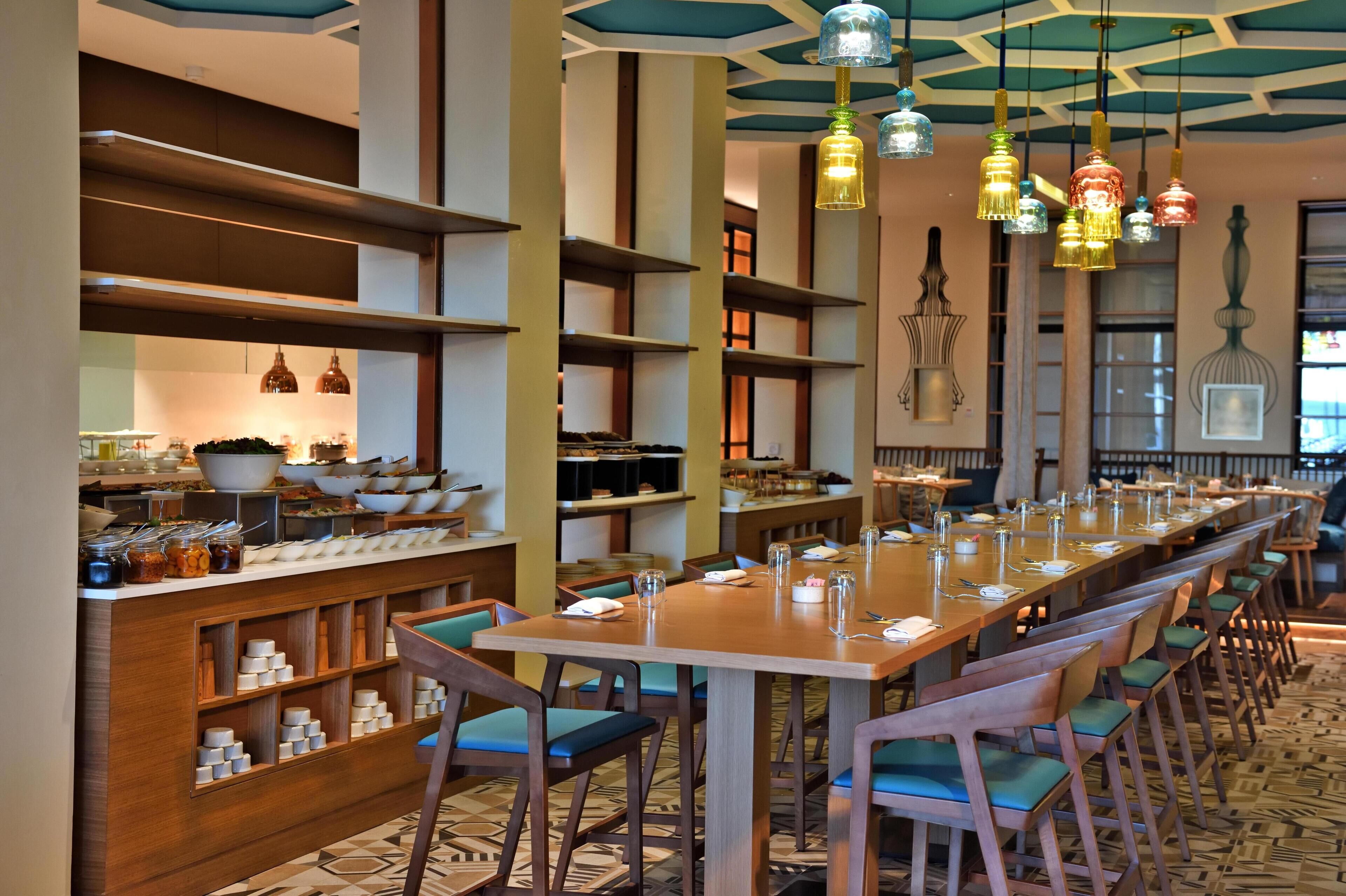 Une longue table à manger entourée de chaises, un espace buffet avec des étagères et des lampes suspendues.