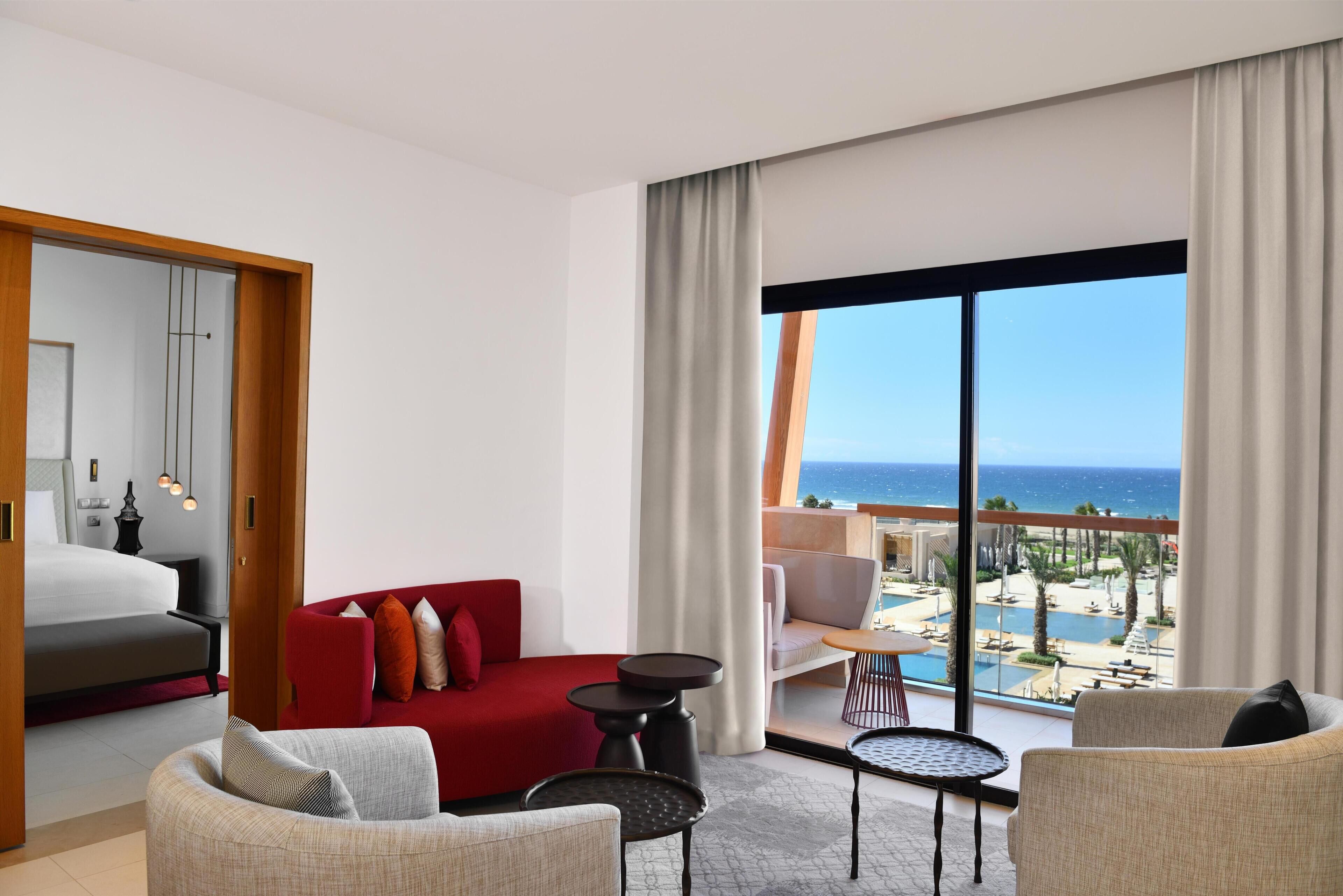 A modern hotel room with a large window, a red sofa, a grey armchair, a small round table, and a bed with a white headboard.