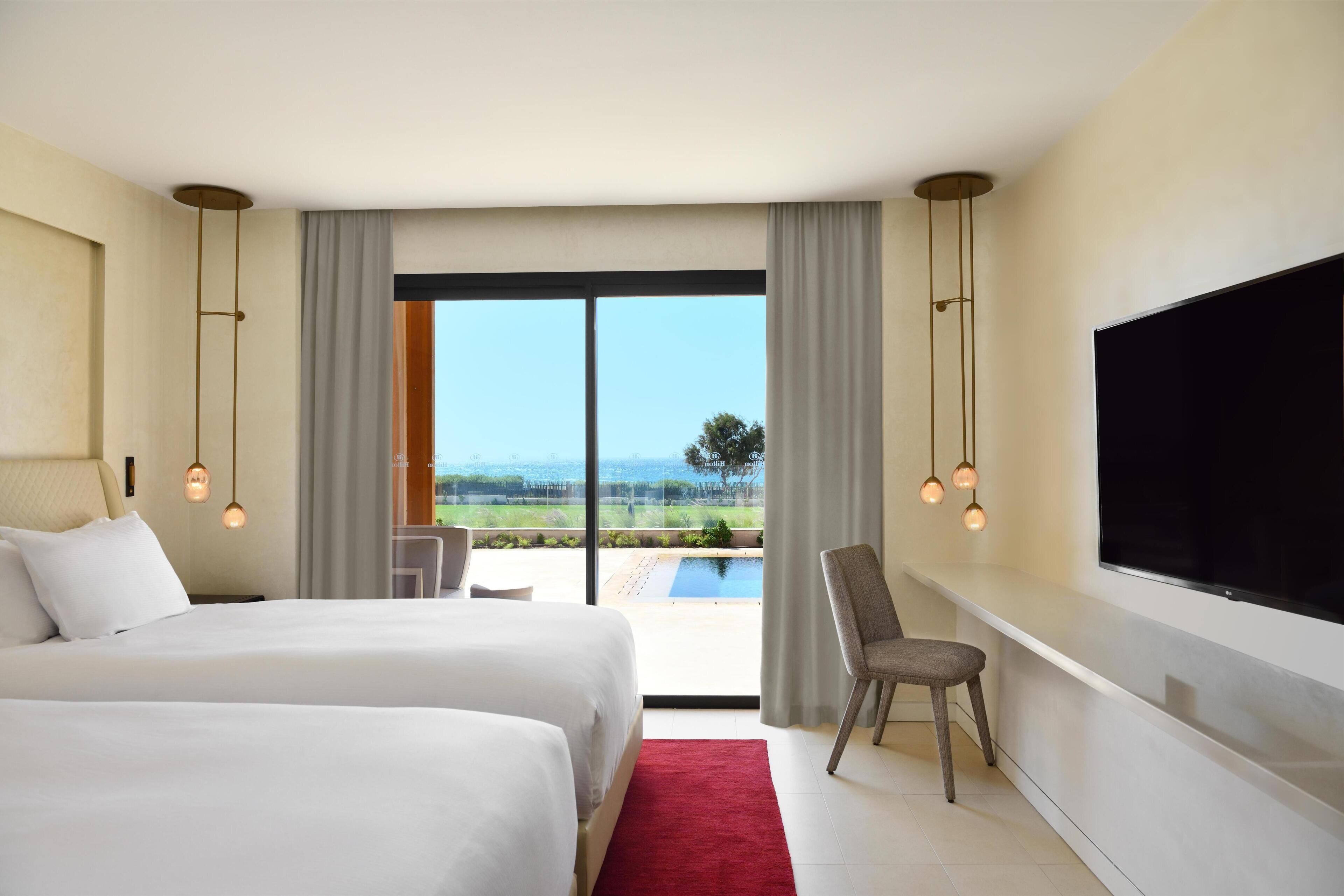 Habitación de hotel con dos camas, televisión, una silla, escritorio y un ventanal con vistas.