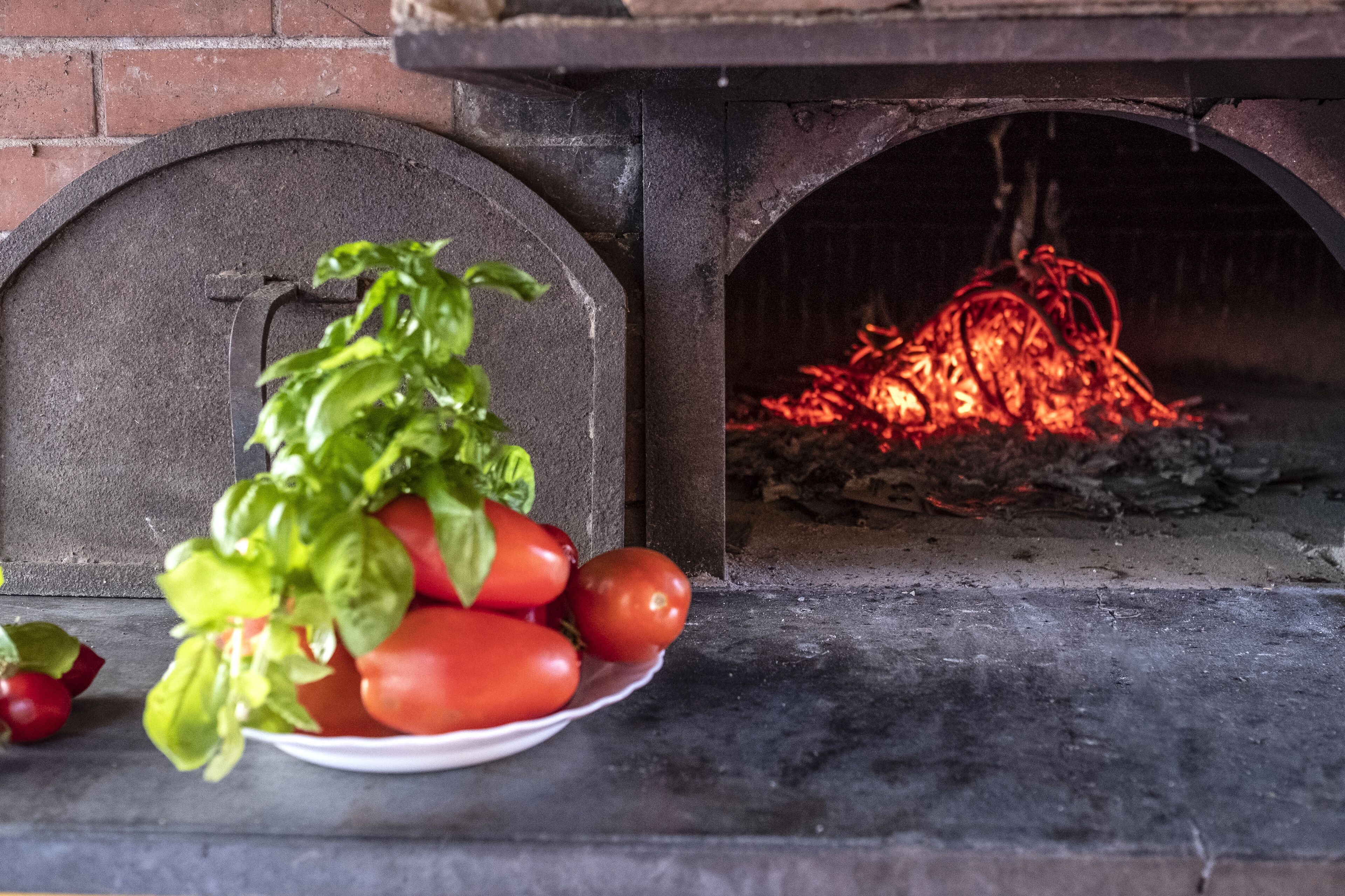 Un four en briques dans lequel brûle du bois, une assiette avec des tomates et des feuilles de basilic, et un mur en briques.