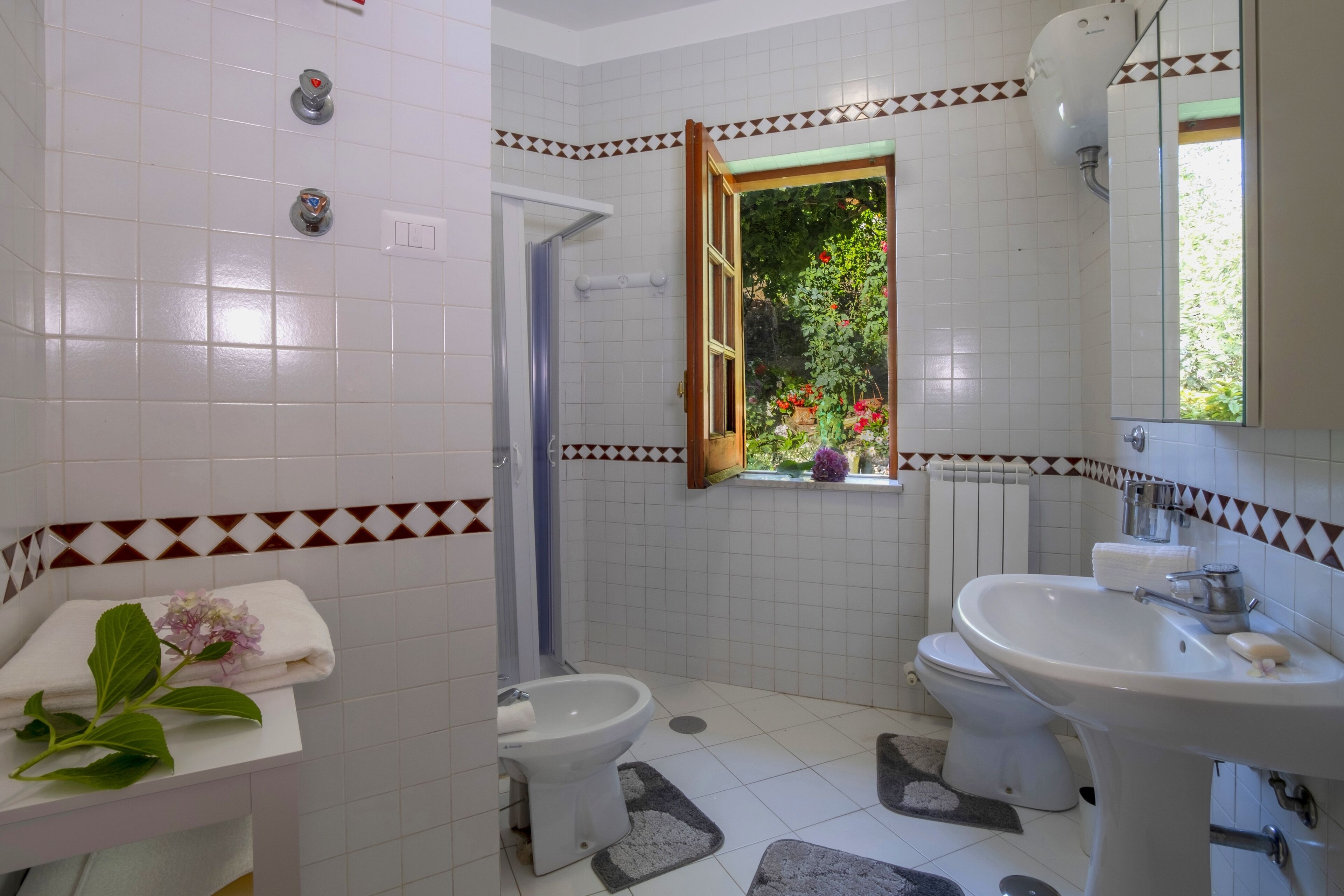 Un bagno con piastrelle bianche, un bidet, un WC, un lavandino e una finestra con vista sul verde.