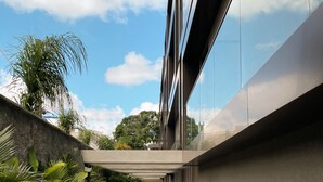Un edificio moderno con fachada de vidrio reflectante, un sendero de concreto bordeado de plantas tropicales y un cielo azul despejado con nubes.