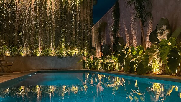 Zona de piscina nocturna con piscina cristalina, plantas circundantes y un edificio con paredes iluminadas.