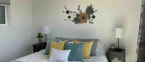 Quarto com cama, duas mesas de cabeceira, ar condicionado, um painel decorativo e janela com persianas.