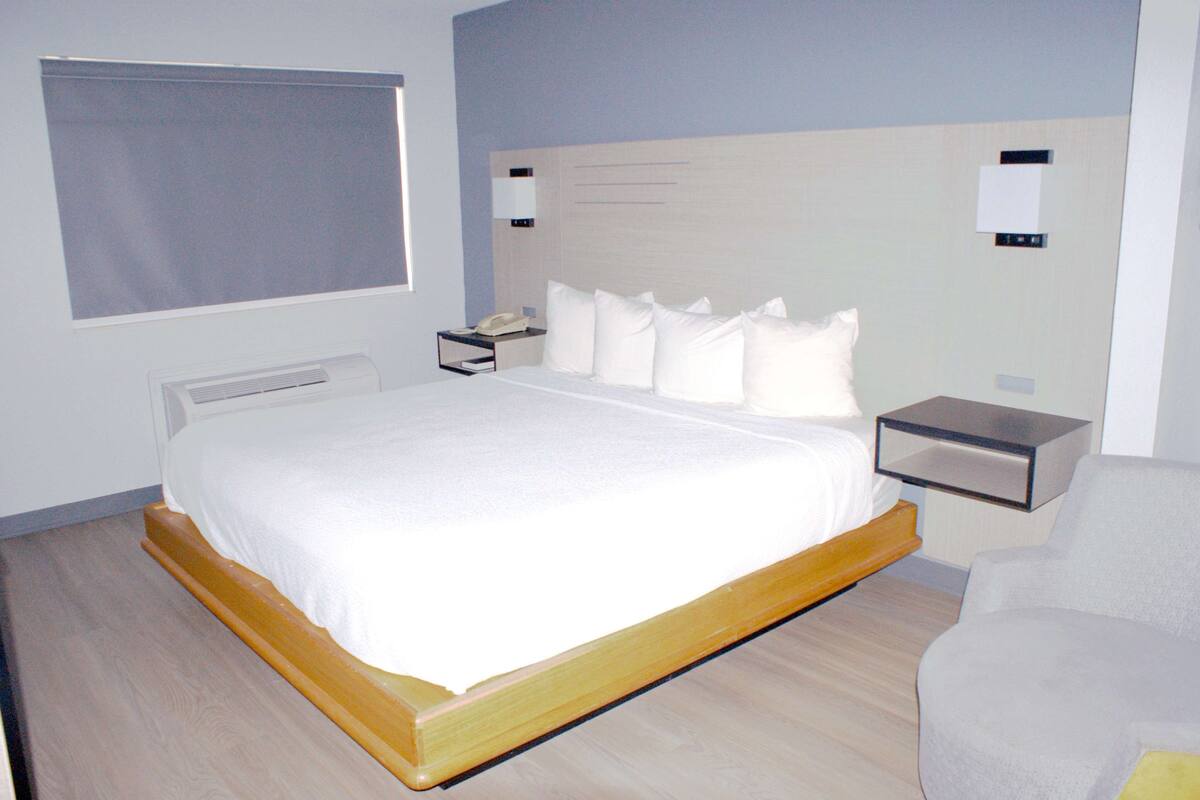 Moderni hotellihuone, jossa on suuri sänky, yöpöytä lampun kanssa, tuoli ja ikkuna, jossa on sälekaihtimet.