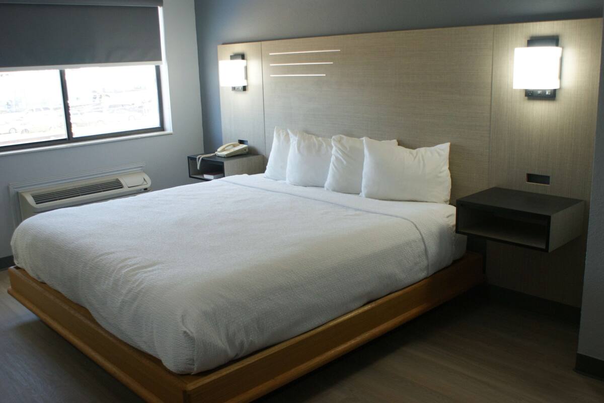 Moderni hotellihuone, jossa on suuri sänky, yöpöydällä olevat lamput, yöpöytä ja ikkuna, jossa on sälekaihtimet.