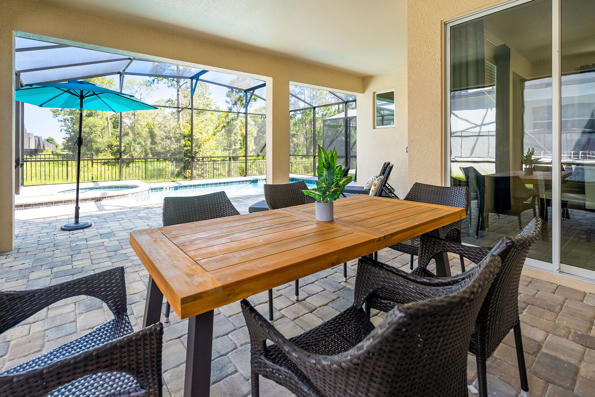 Un patio al aire libre con una mesa de madera y sillas de mimbre, una planta en maceta y vista a una piscina.