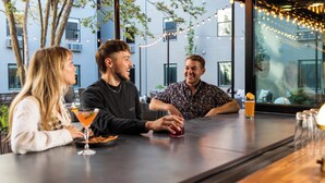 Drie personen zitten aan een buitenbar, versierd met lichtslingers, met een glas jus d'orange en een martini.