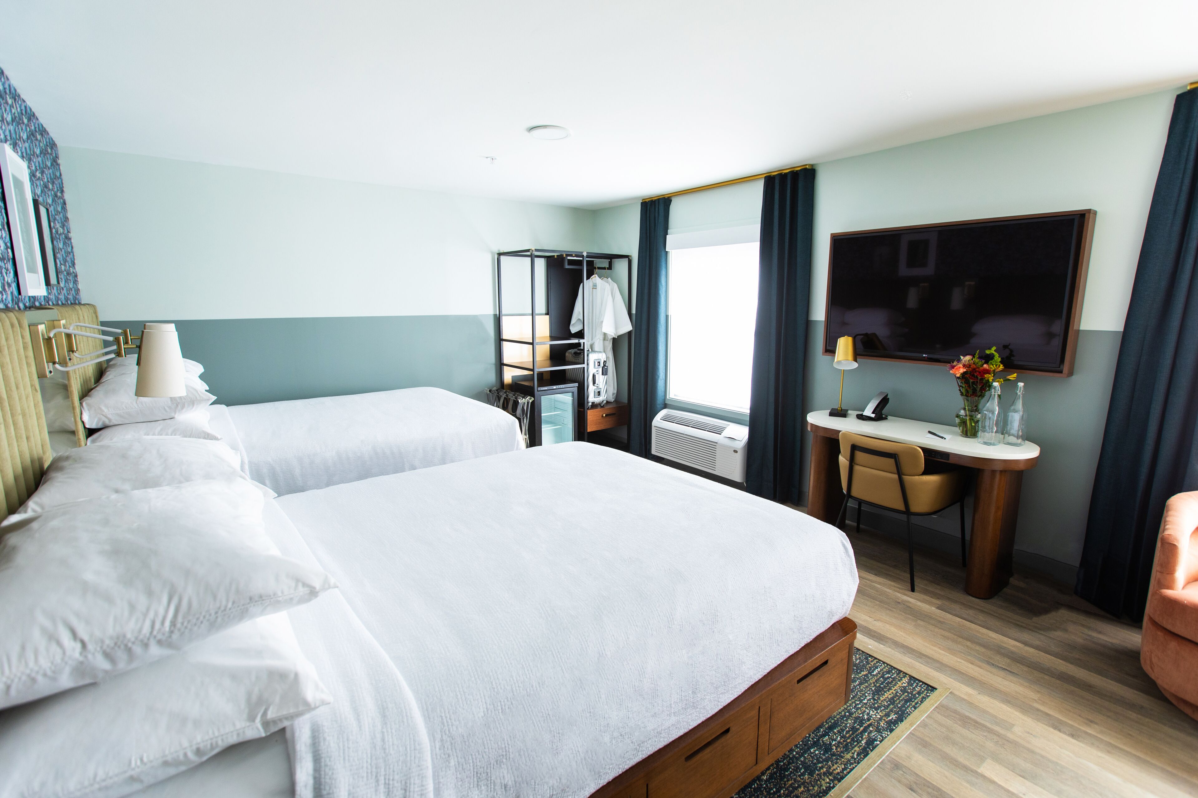 Quarto de hotel com duas camas, uma escrivaninha, uma cadeira, uma televisão e uma janela com cortinas.