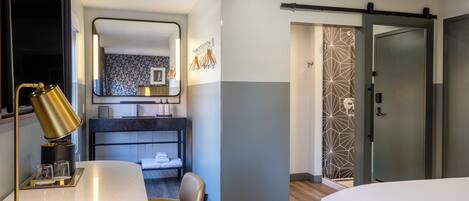A modern hotel room with a bed, a desk with a lamp, a chair, a bathroom with a shower, and a sliding door.