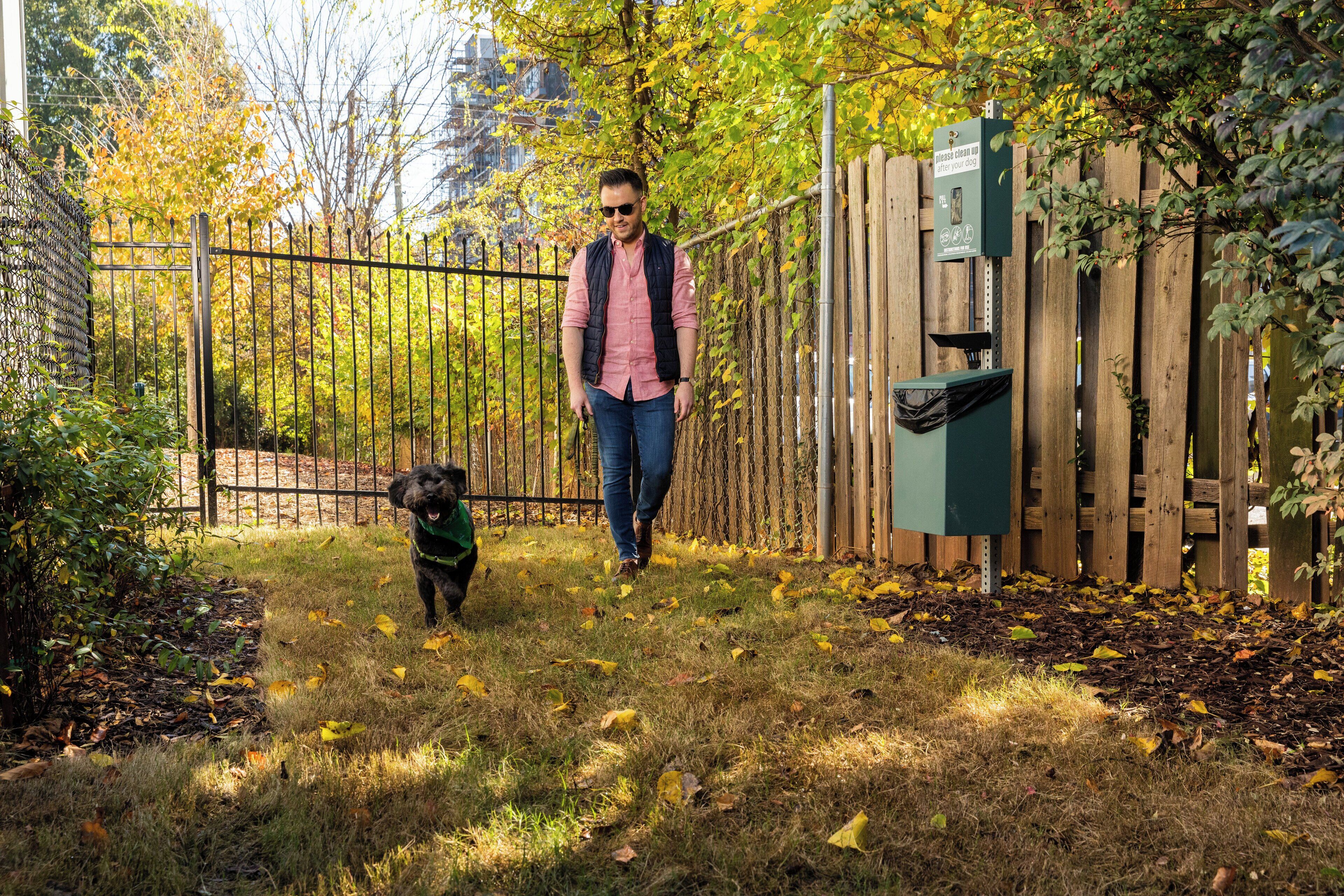 Un home passeja un gos per un pati tancat amb una valla de fusta i un cubell de color verd.
