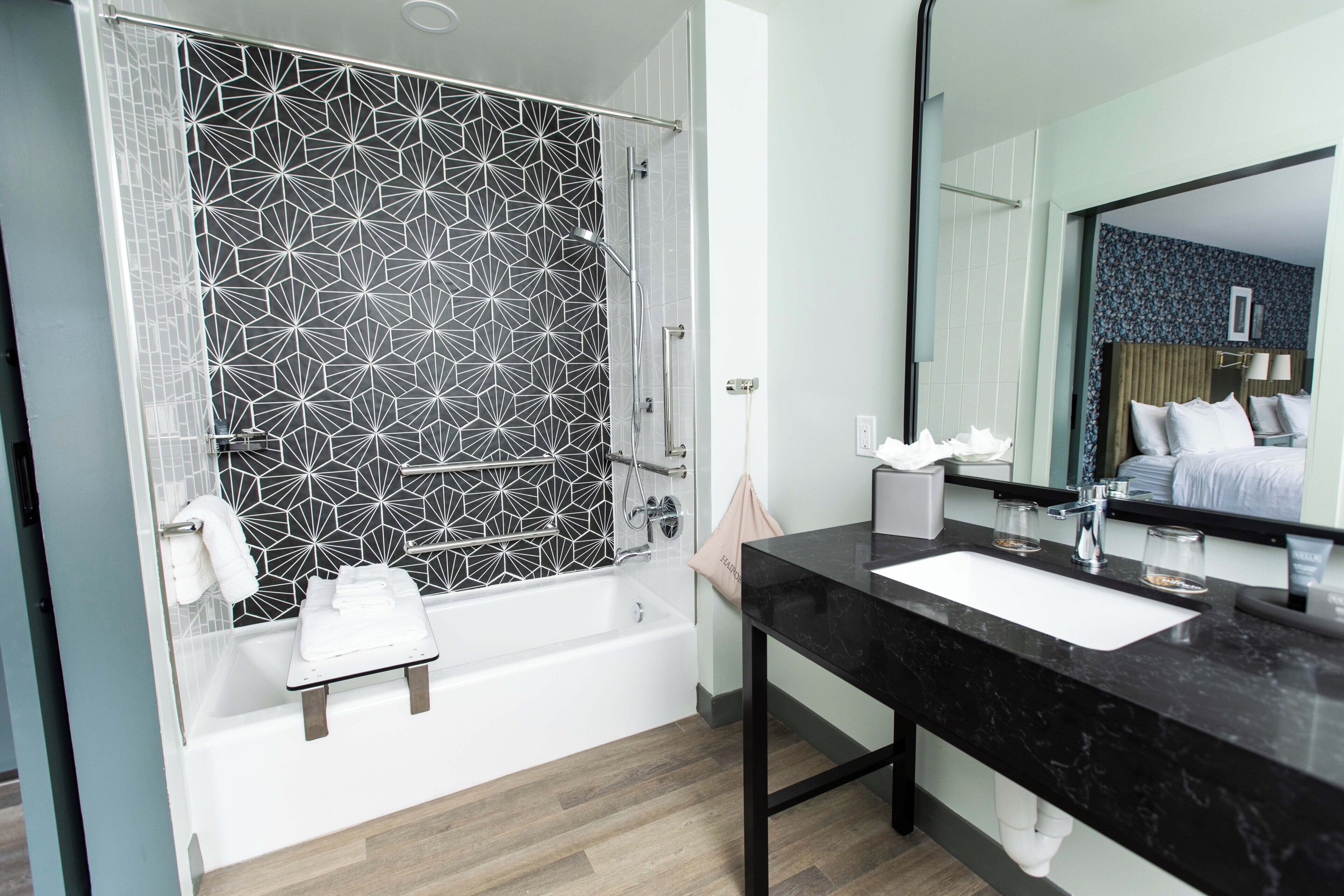 A modern bathroom with a geometric-patterned shower enclosure, a white bathtub, a black marble sink, and a large mirror reflecting a bedroom with a bed and a nightstand.