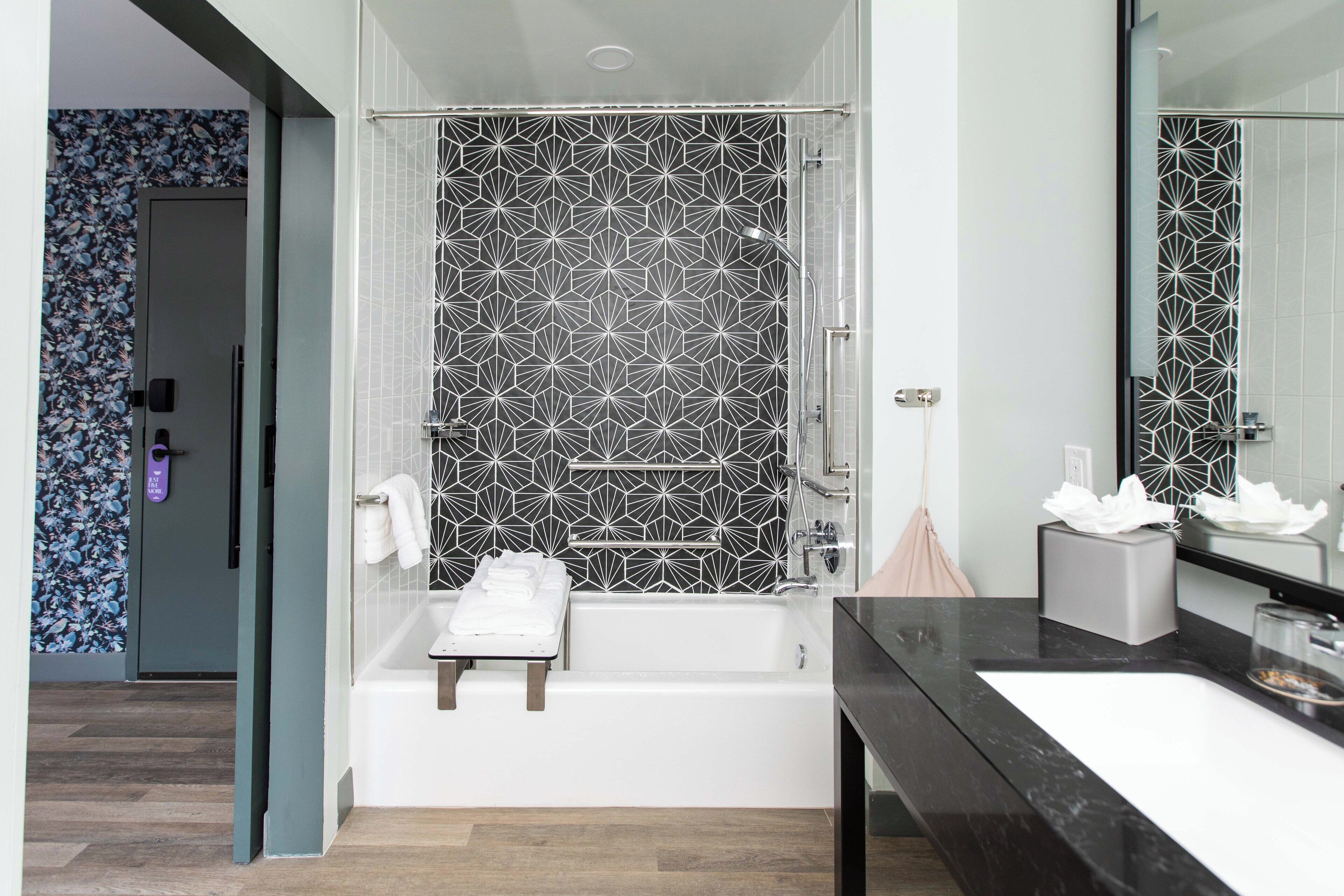 Un baño moderno con pared de ducha de azulejos geométricos, una bañera blanca y un lavabo sobre encimera negra.