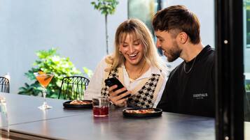 Una pareja sentada a una mesa, con una bebida y un plato de comida, en una habitación con un ventanal grande y una planta afuera.