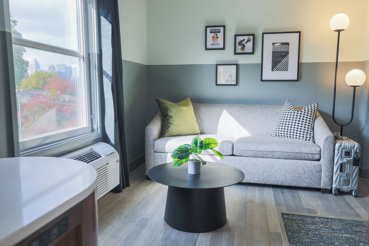 A modern living room with a grey sofa, a round black coffee table, and a potted plant. There are framed pictures on the wall, a standing lamp, and a window with a view of trees.