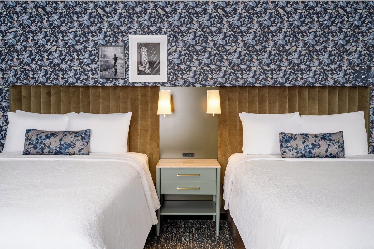 A hotel room with two beds, a wooden headboard, a nightstand, and a floral-patterned wallpaper.