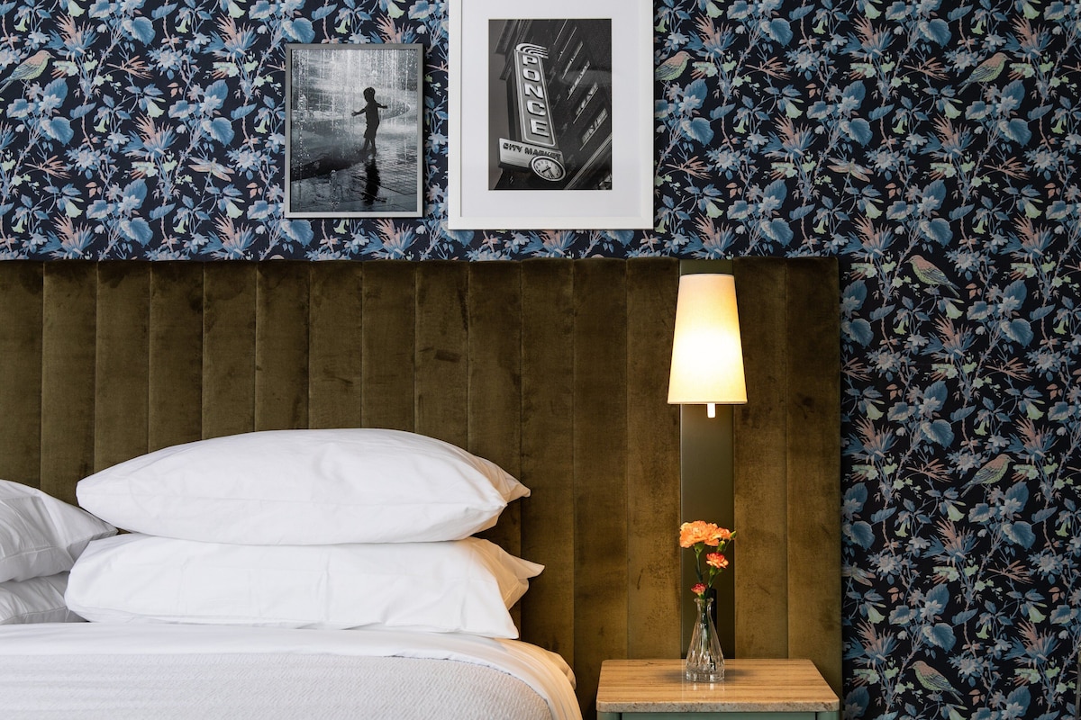 A neatly made bed with white linens, a wooden headboard, a nightstand with a lamp, and a framed picture on the wall.