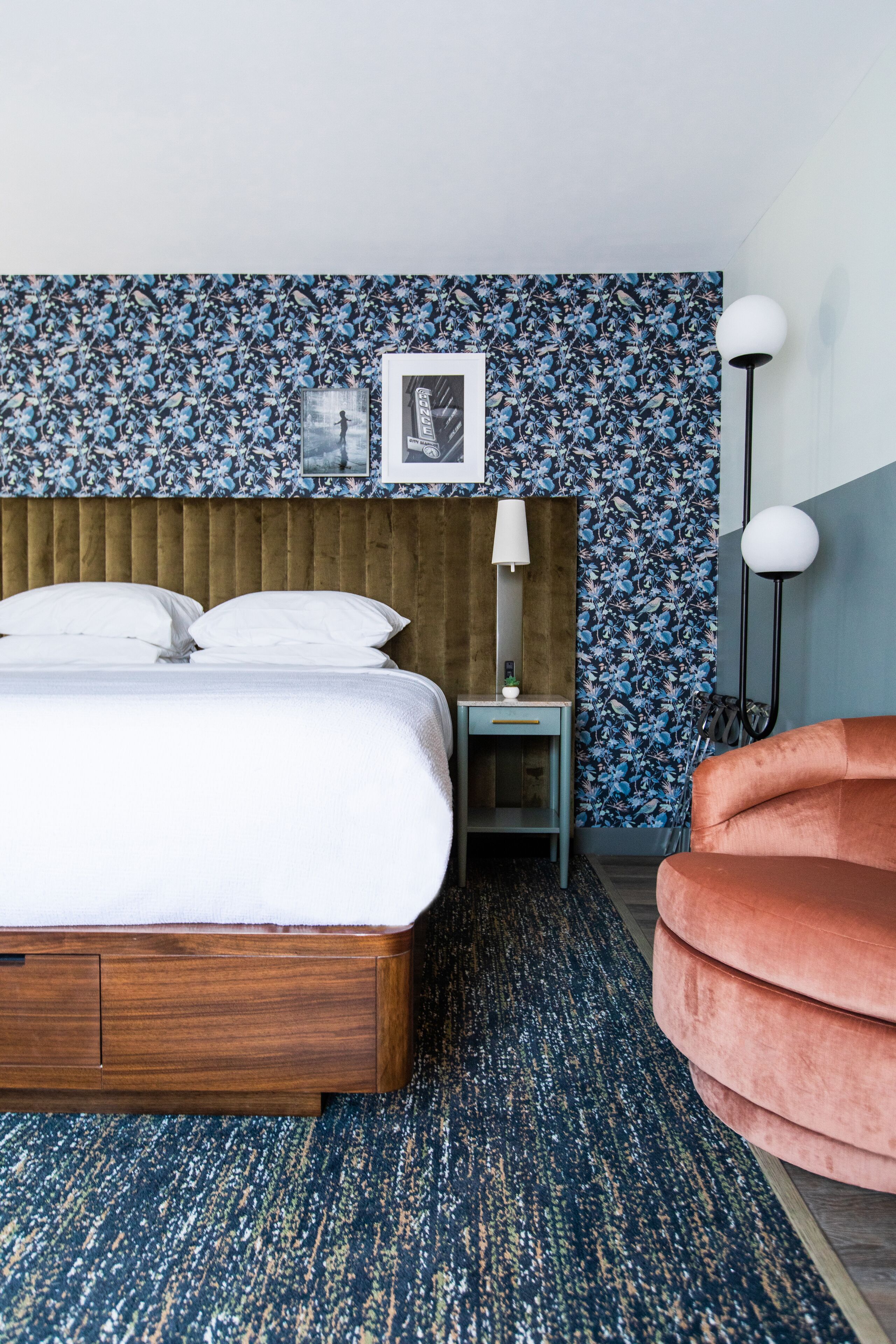 A hotel room with a wooden bed, a nightstand with a lamp, a chair, and a wall with a floral pattern.