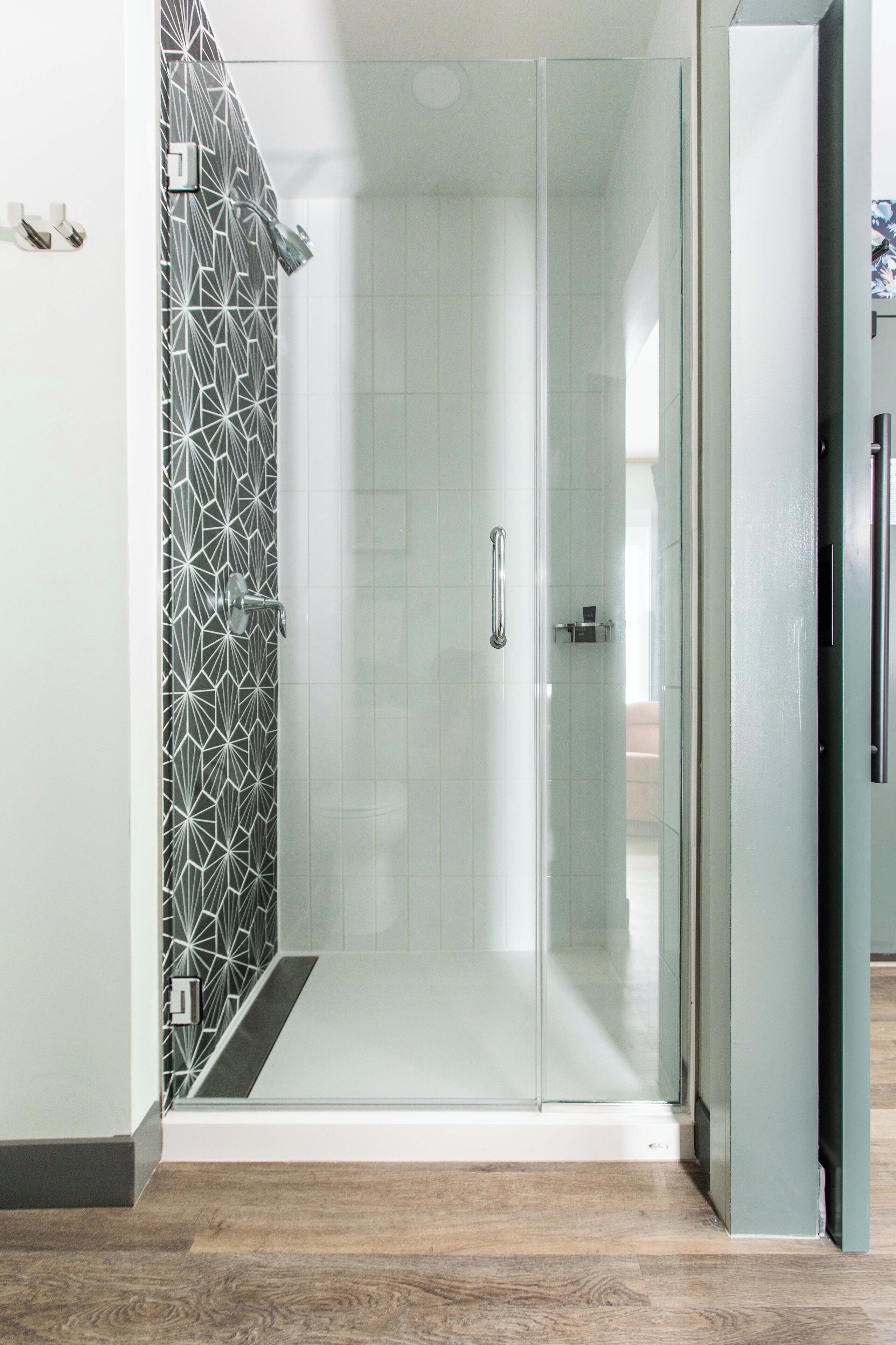 A modern shower area with a glass door, geometric wall pattern, and a wooden floor.