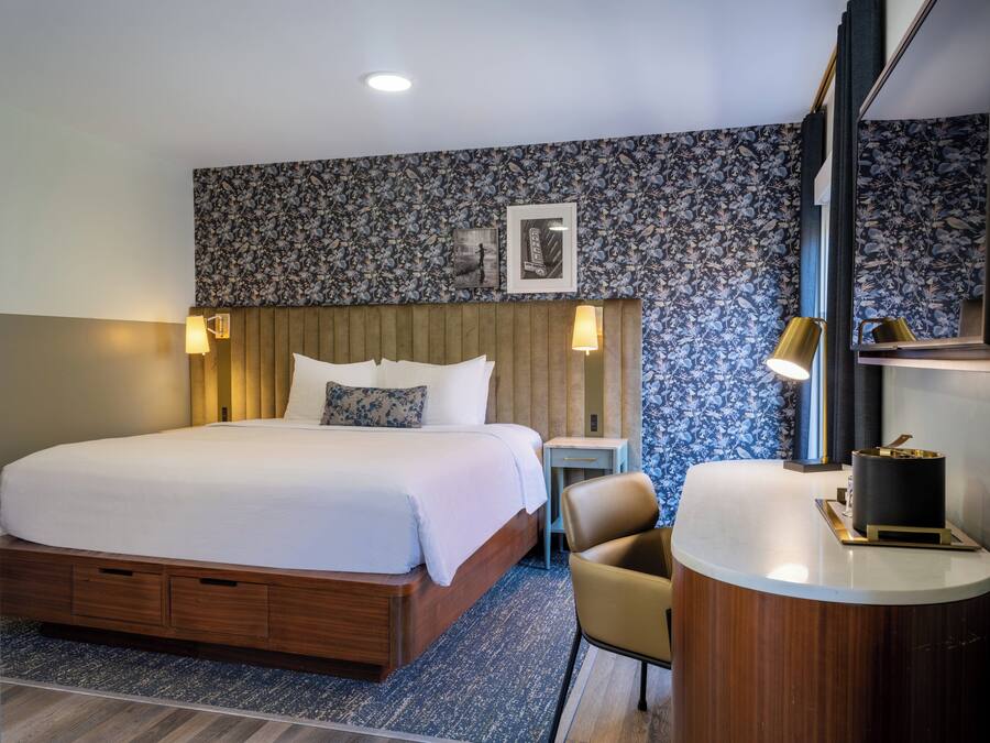 Habitación de hotel con una cama grande, cabecera de madera, un escritorio pequeño con una silla, una mesa redonda y un espejo.
