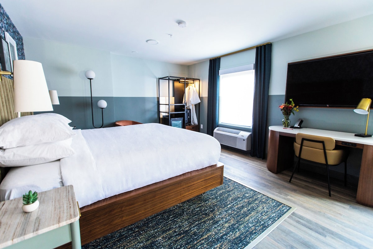 A hotel room with a bed, desk, chair, TV, and a window with curtains.