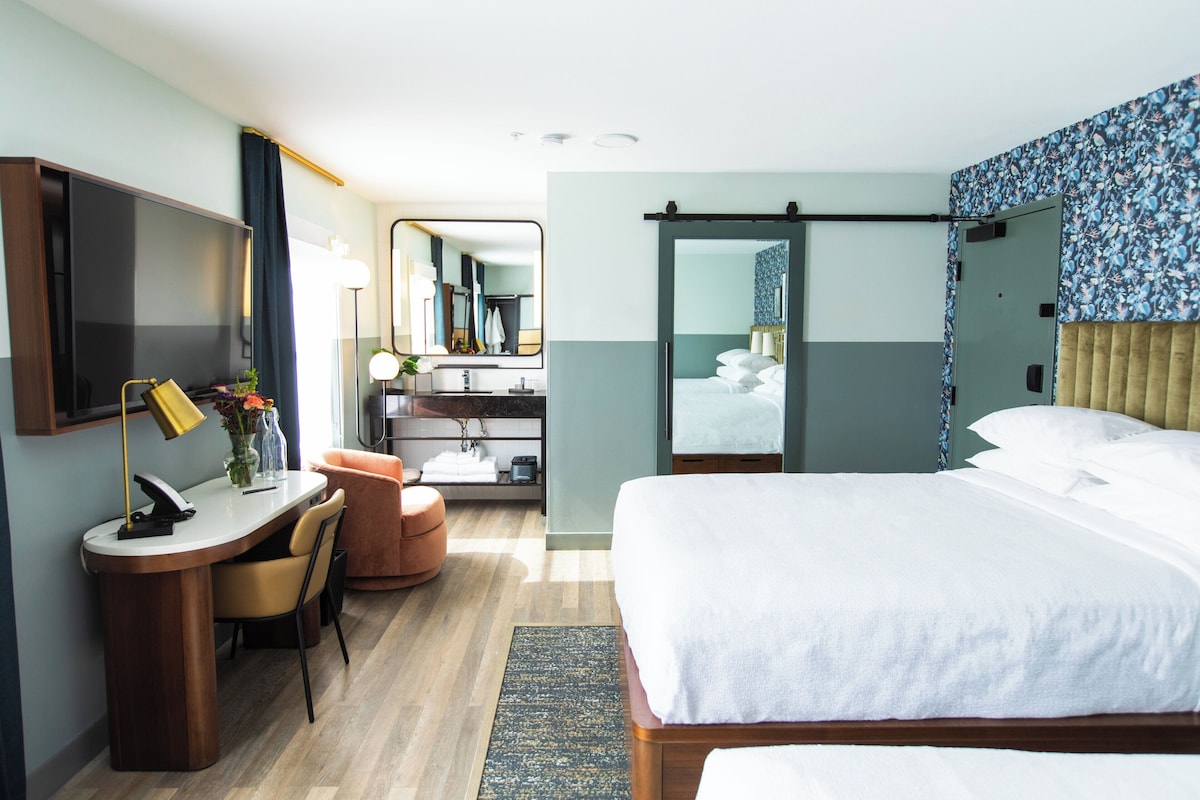 Habitación de hotel con cama, escritorio con silla, espejo, baño con lavamanos y espejo, y zona de ducha.