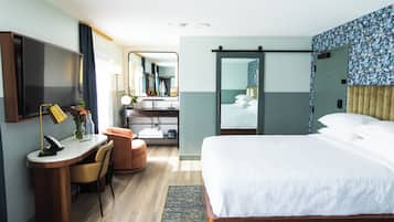 A hotel room with a bed, a desk with a chair, a mirror, a bathroom with a sink and mirror, and a shower area.