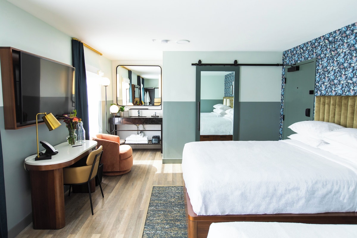 A hotel room with a bed, a desk with a chair, a mirror, a bathroom with a sink and mirror, and a shower area.