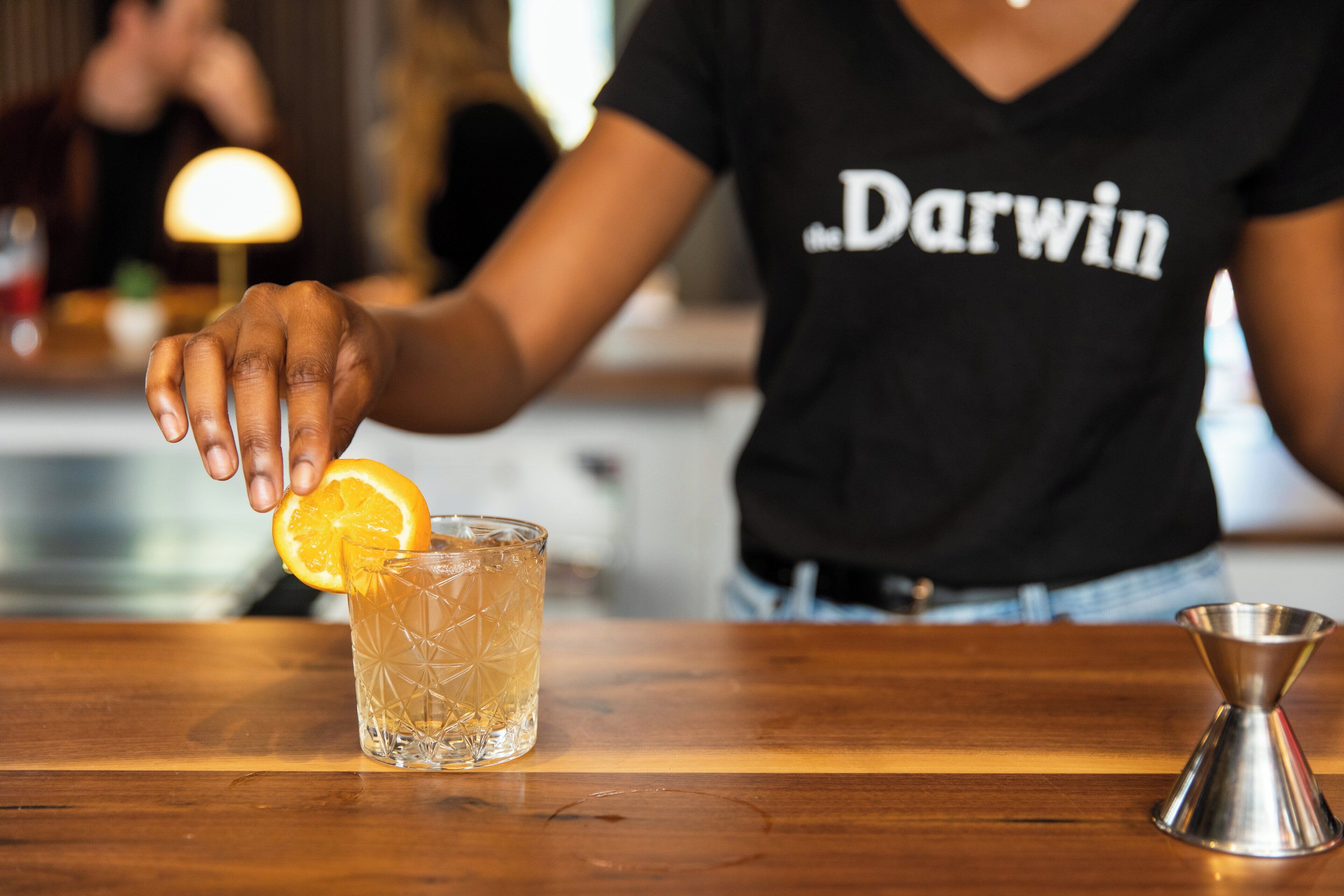 A person in a black shirt with "the Darwin" text, placing an orange slice on a clear glass with a geometric pattern. A metal shaker is on the table beside the glass.