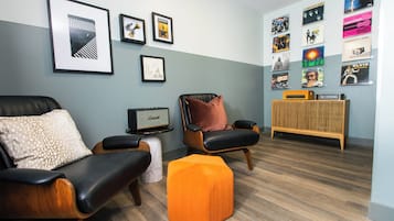 Un salon moderne avec un canapé, un pouf et une table basse. Des tableaux encadrés ornent le mur, et une petite table porte une radio.