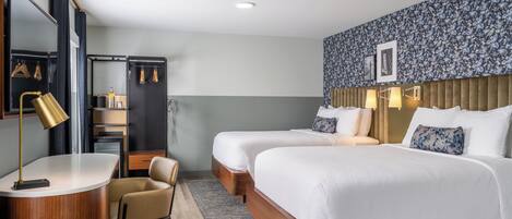 A hotel room with two beds, a desk with a lamp, a chair, a wardrobe, and a shower area.