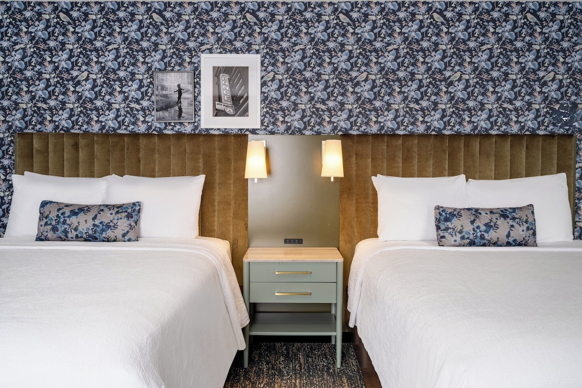 Habitación de hotel con dos camas, cabecero de madera, mesita de noche y papel tapiz con estampado floral.