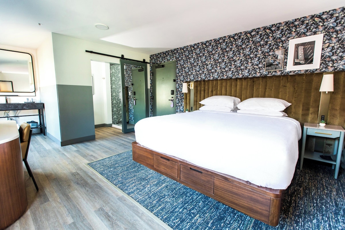 Una habitación de hotel moderna con una cama grande, un escritorio con una silla, un vestidor y un baño con ducha e inodoro.