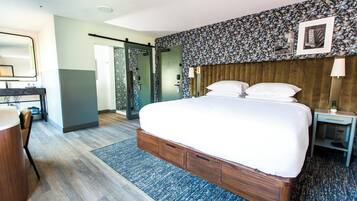 A modern hotel room with a large bed, a desk with a chair, a walk-in closet, and a bathroom with a shower and toilet.
