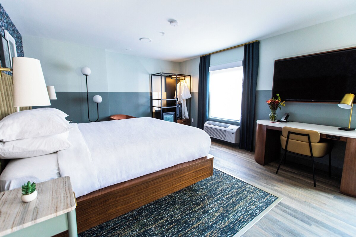 Una habitación de hotel con cama, escritorio, silla, televisión y ventana con cortinas.