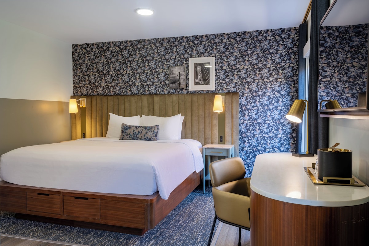 A hotel room with a large bed, a wooden headboard, a small desk with a chair, a round table, and a mirror.