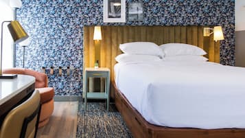 Ett hotellrum med en sänggavel i trä, en säng med vita sängkläder, ett litet nattduksbord med en lampa, en stol och ett skrivbord.