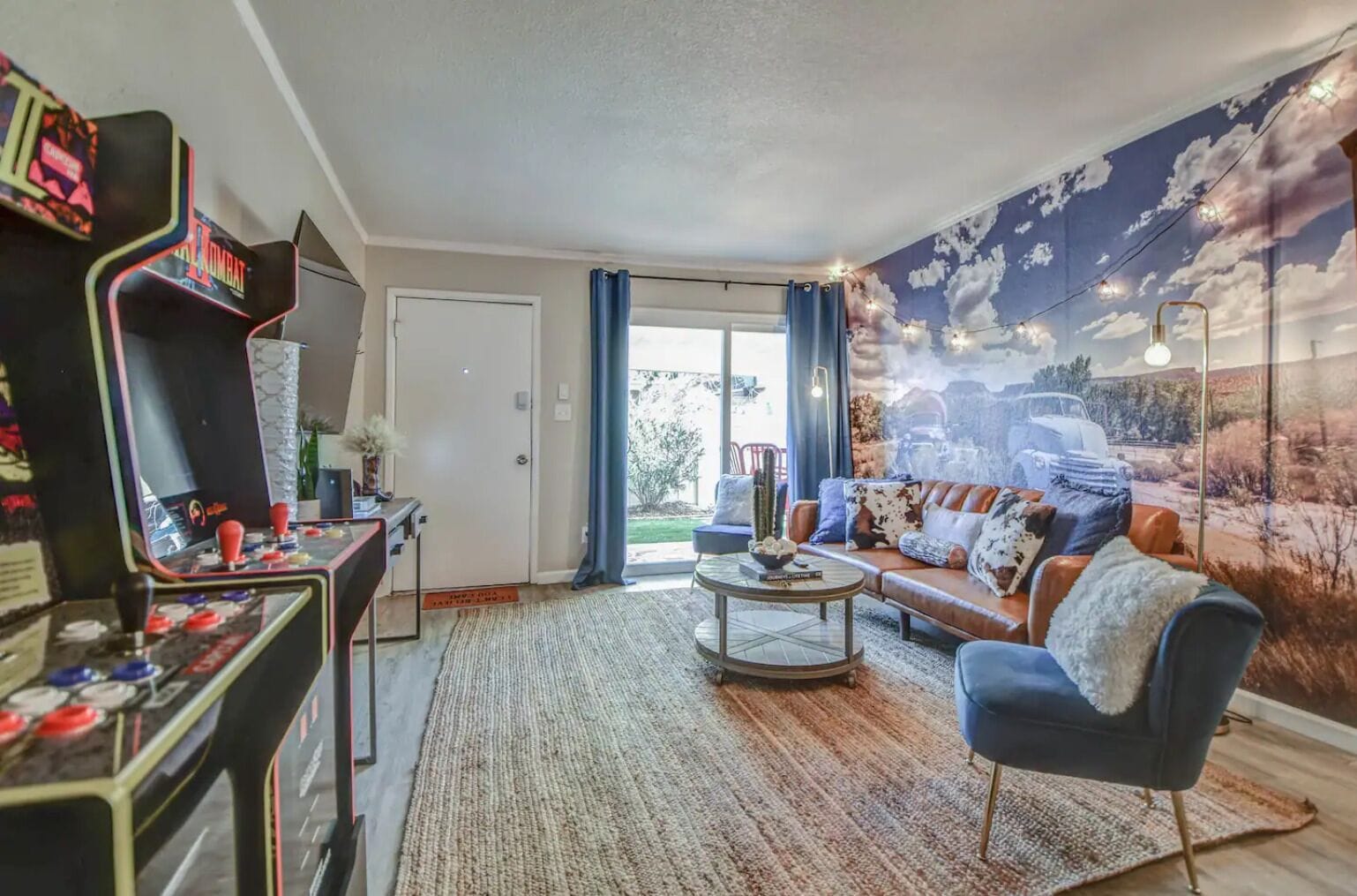Een woonkamer met een retro-arcadekast, een bank met gestreepte kussens, een bijzettafeltje en een muurschildering van een auto in een woestijnlandschap.