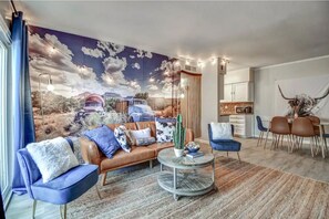 Ein Wohnzimmer mit Wandgemälde einer Wüstenlandschaft, einem braunen Ledersofa, blauen Sessel und einem runden Couchtisch. Zusätzlich ein Essbereich mit weißem Tisch und braunen Stühlen.