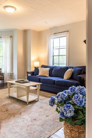 A living room with a blue sofa, a coffee table, a lamp, and a window with curtains. - Raspberry Inn · Classic Riverfront Home-Mins to Woodstock & Skiing (Bridgewater)