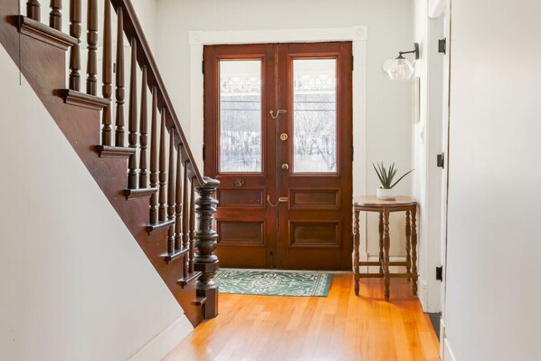 A wooden staircase leading to a double door with a glass panel, a small round table with a plant, and a green doormat.
